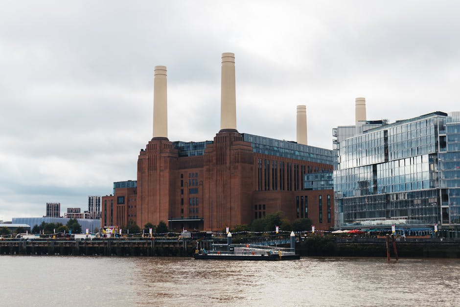 A large industrial building with a distinctive red brick facade and four tall white chimneys extending upward, situated along the riverbank with the water visible in the foreground. The building is ad
