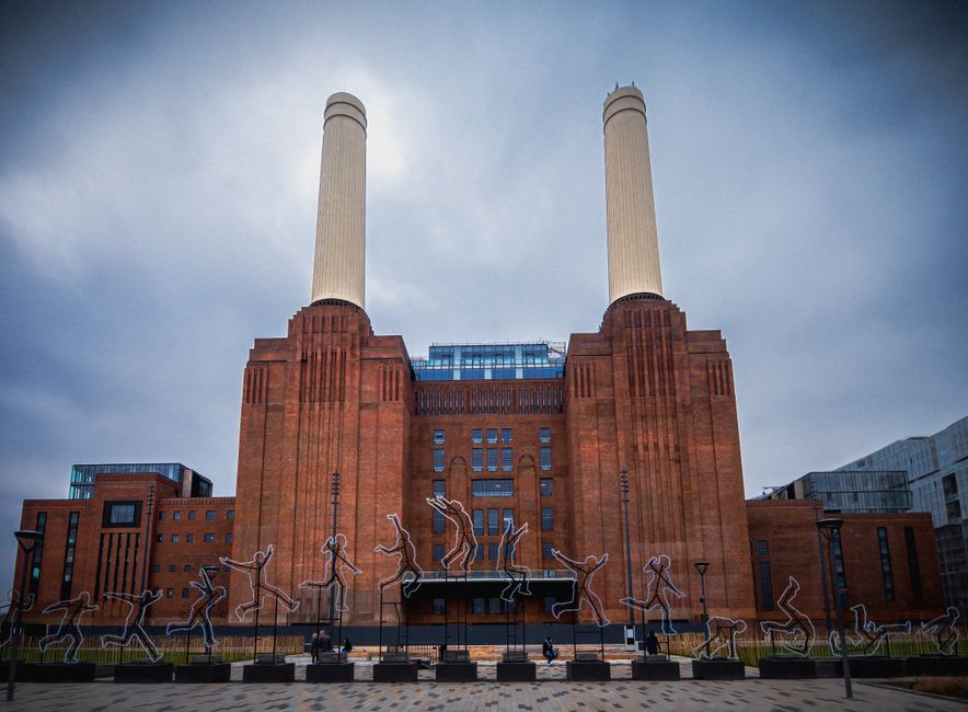 View of the iconic Battersea Power Station with its distinctive four tall chimneys, situated along the River Thames on a cloudy day. In the foreground, the river water appears calm with reflections of