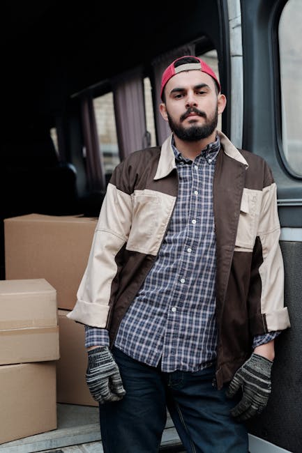 A male removal worker with a beard, wearing a red cap, a checkered shirt, and a two-tone beige and brown jacket, standing inside a van during a home relocation process. The worker is wearing grey glov